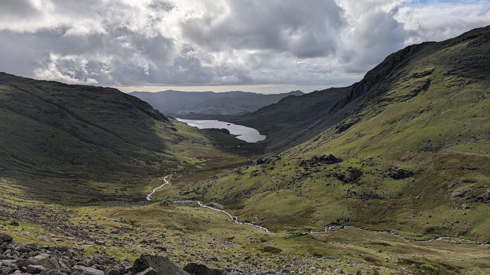 A stream winds down a green valley towards a tarn, under heavy cloud